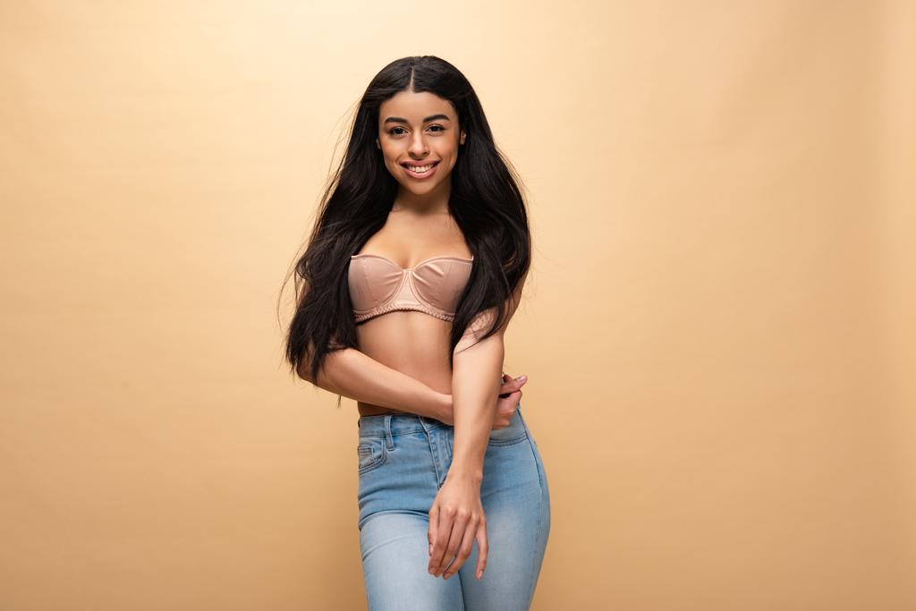 pretty african american girl in blue jeans and bra posing at camera isolated on beige  - Photo, Image
