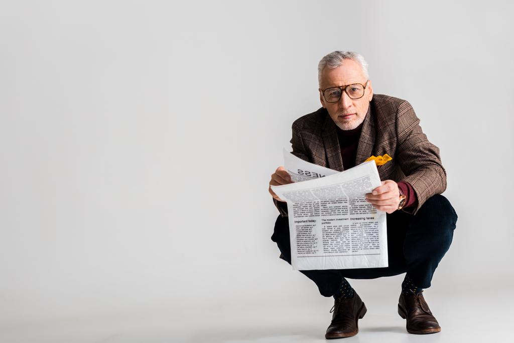Трехлетний взрослый мужчина в очках, держа в руках газету, пока сидит на сером
 - Фото, изображение