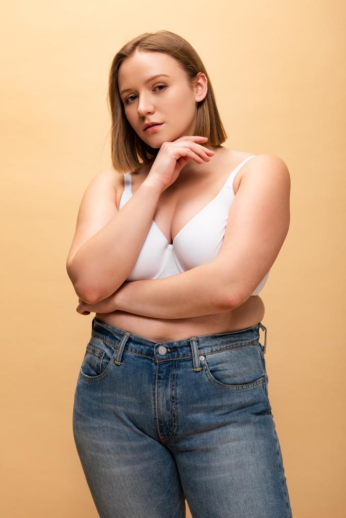 pensive overweight girl looking at camera while holding hand near face isolated on beige, body positivity concept - Photo, Image