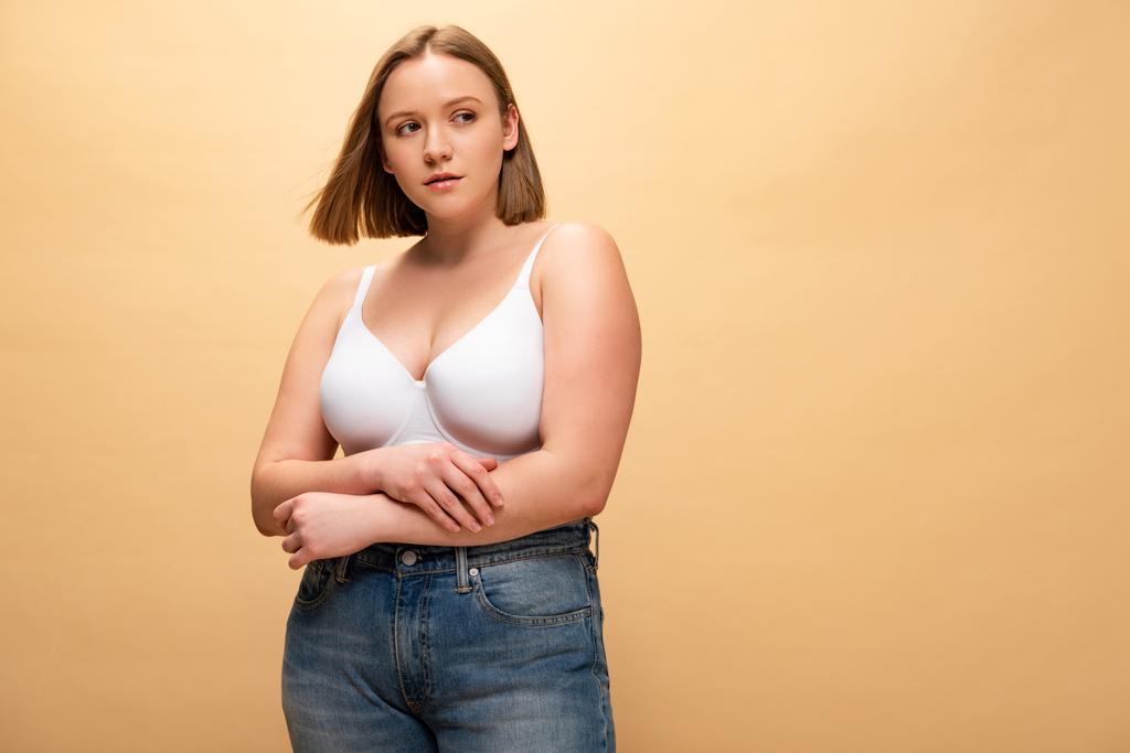 mietteliäs ylipainoinen tyttö sinisissä farkuissa ja rintaliiveissä etsii pois eristettynä beige, kehon positiivisuus käsite
 - Valokuva, kuva
