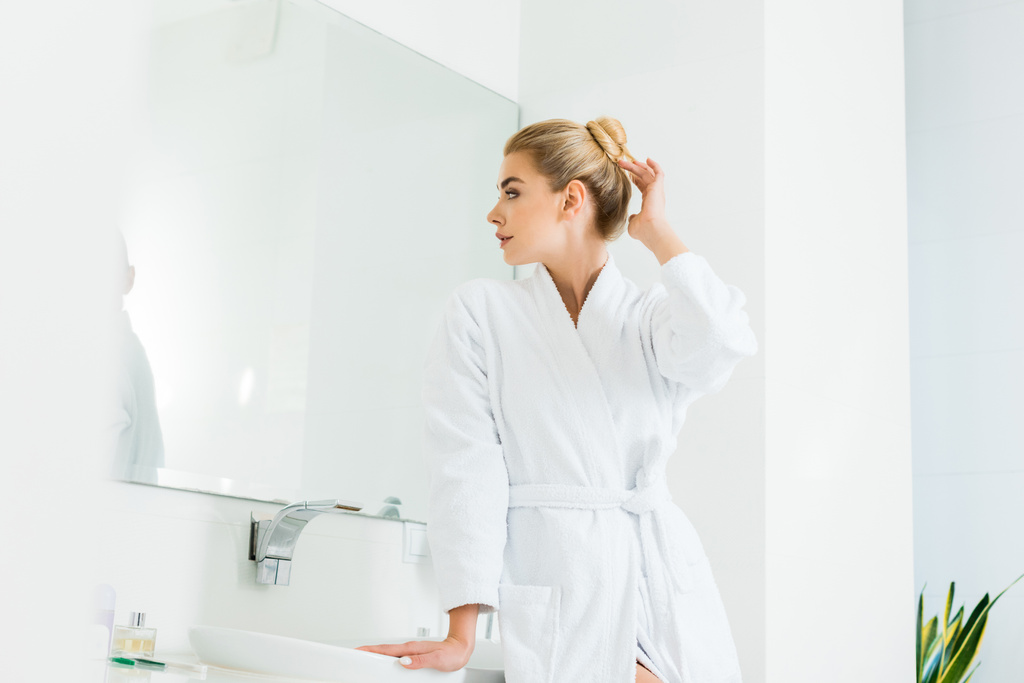 attraktive und blonde Frau im weißen Bademantel, die im Badezimmer in den Spiegel schaut  - Foto, Bild