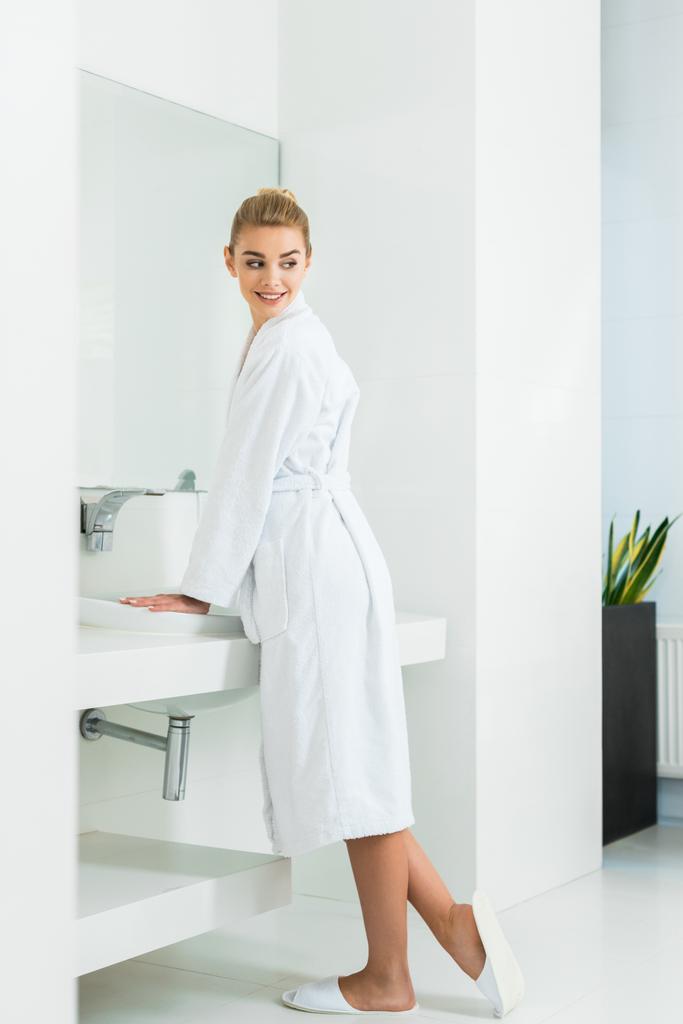 schöne und lächelnde Frau im weißen Bademantel und Hausschuhen, die im Badezimmer wegschaut  - Foto, Bild