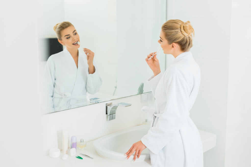 Selektiver Fokus der Frau im weißen Bademantel beim Zähneputzen und Blick in den Spiegel  - Foto, Bild