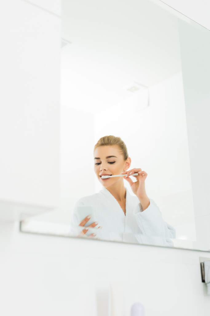 γυναίκα στο λευκό μπουρνούζι βούρτσισμα των δοντιών και η χρήση smartphone στο μπάνιο  - Φωτογραφία, εικόνα