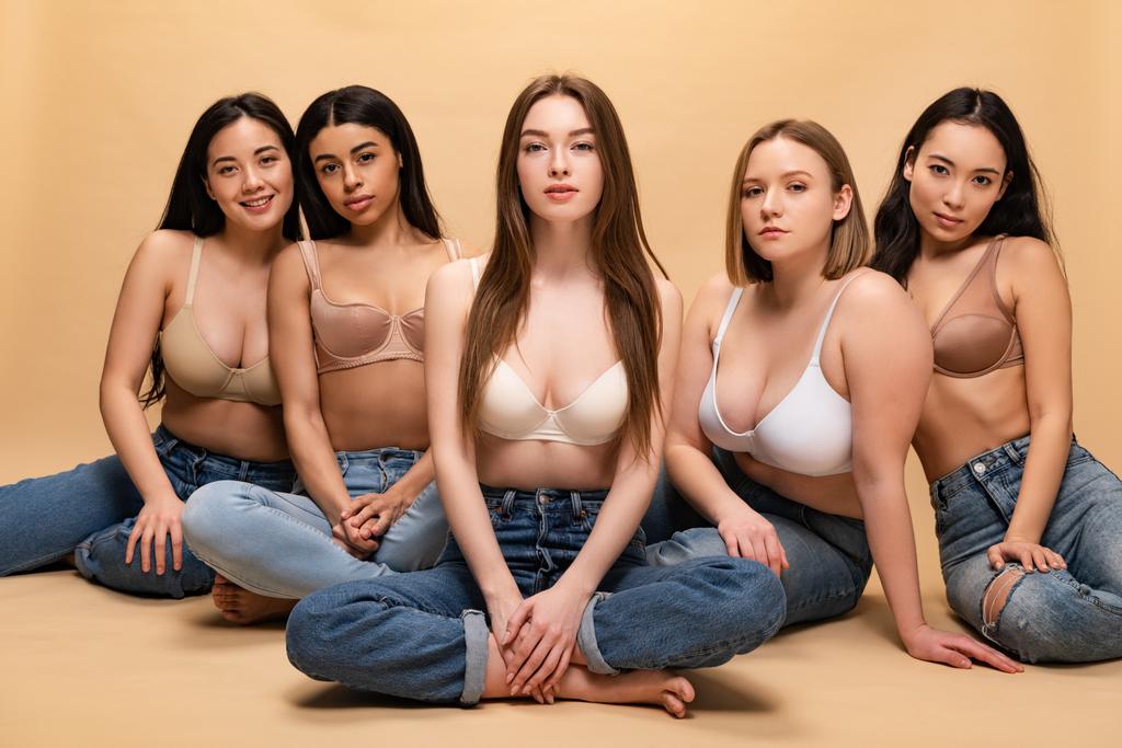 пять красивых мультикультурных девушек в синих джинсах и бюстгальтерах, сидящих и смотрящих на камеру, концепция позитивности тела
 - Фото, изображение