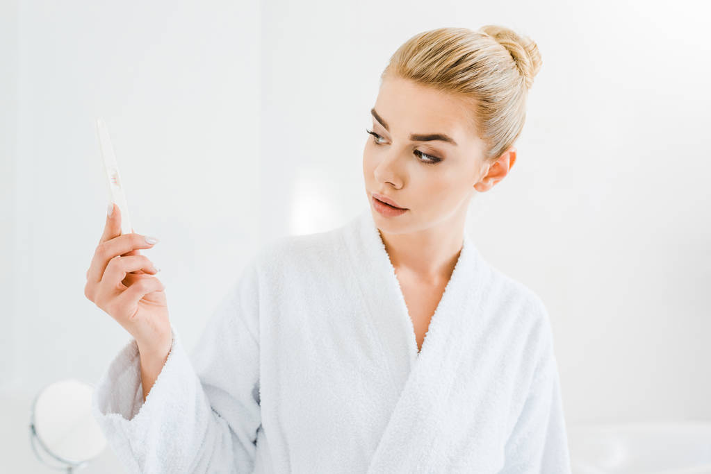 atrakcyjny i blond kobieta w biały szlafrok gospodarstwa pilnik w łazience  - Zdjęcie, obraz