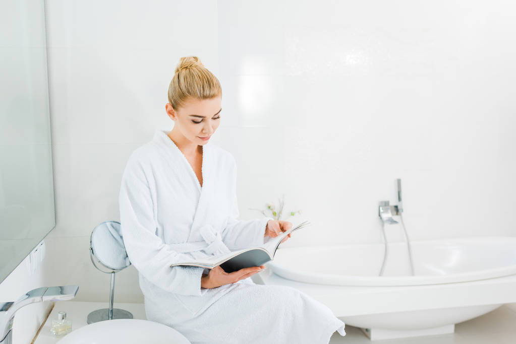 femme attrayante et blonde en peignoir blanc lecture magazine dans la salle de bain
  - Photo, image