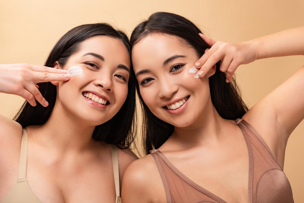 deux femmes multiculturelles heureuses appliquant de la crème cosmétique sur le visage et regardant la caméra isolée sur beige
 - Photo, image