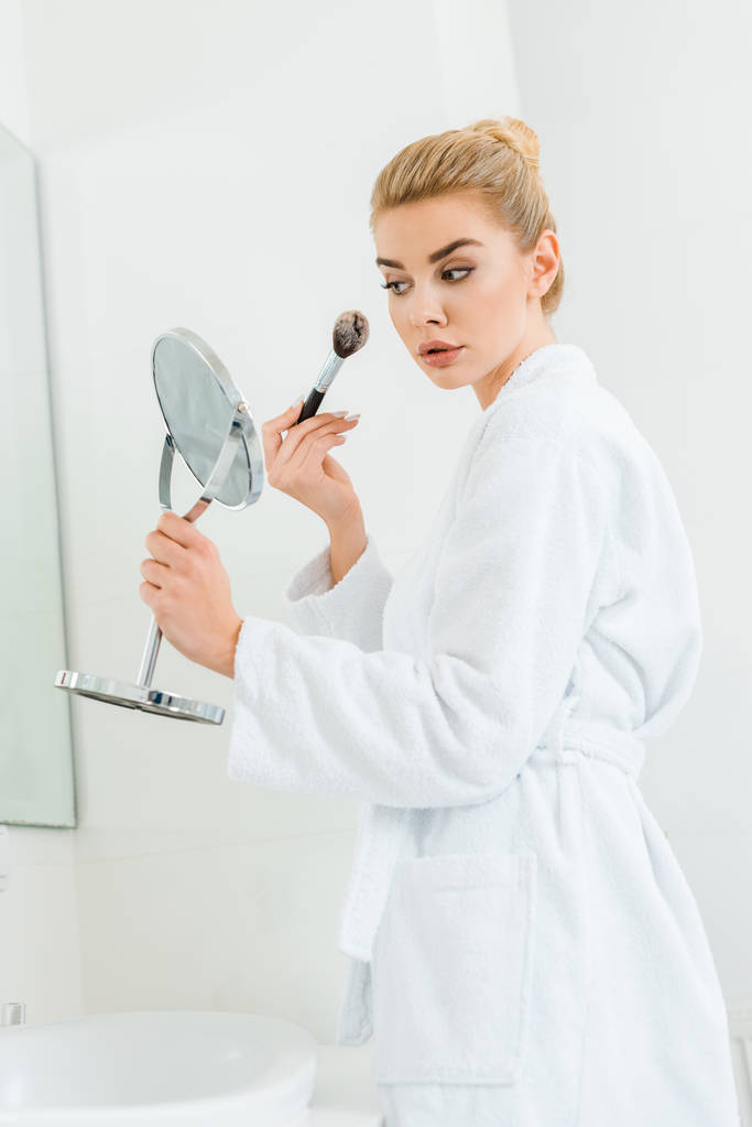 привлекательная блондинка в белом халате с использованием косметической щетки в ванной комнате
  - Фото, изображение