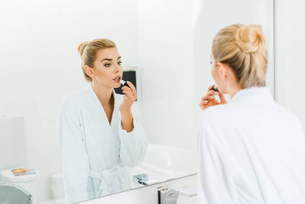enfoque selectivo de la mujer en albornoz blanco aplicando lápiz labial y mirando el espejo en el baño
  - Foto, Imagen