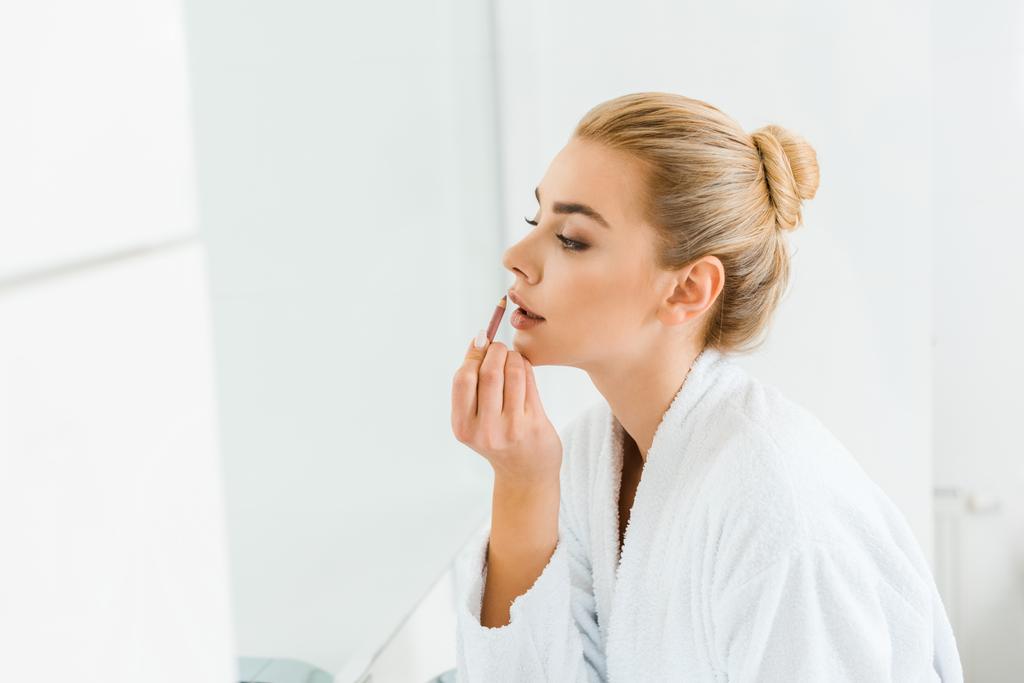 mulher bonita e loira em roupão branco aplicando forro labial no banheiro
  - Foto, Imagem