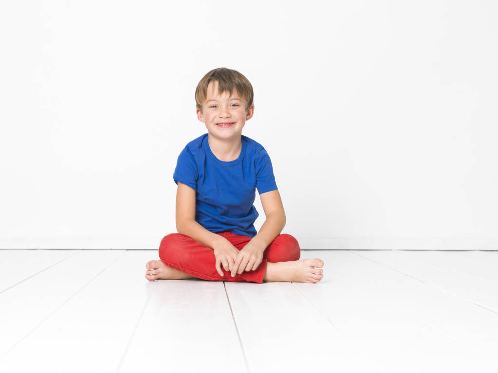 niño de seis años en pantalones rojos y camiseta azul sonriendo y mirando a la cámara mientras está sentado con las piernas cruzadas en el suelo blanco
 - Foto, imagen