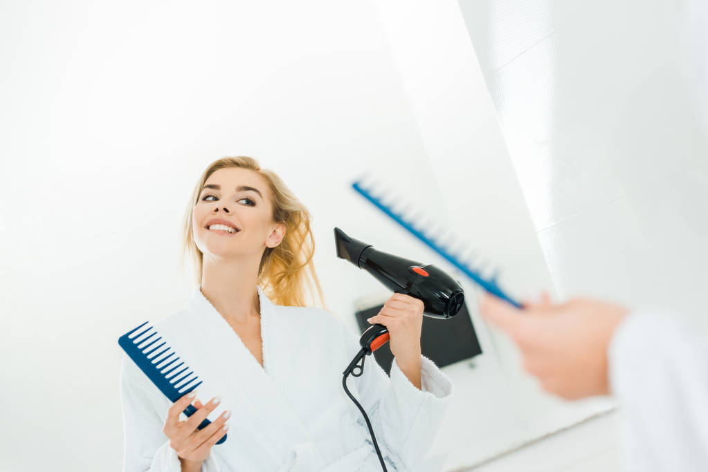 saç kurutma makinesi ve tarak holding beyaz bornoz içinde güzel ve gülümseyen kadın seçici odak - Fotoğraf, Görsel