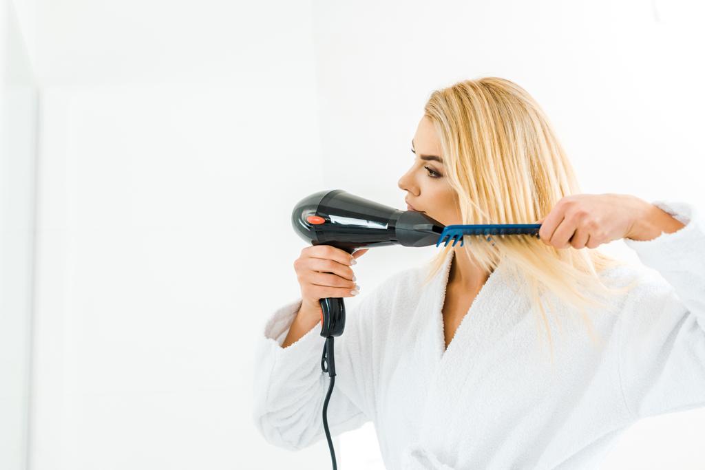 beautiful and blonde woman in white bathrobe using hairdryer and comb - Photo, Image