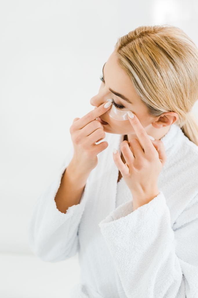 donna bionda e attraente in accappatoio bianco applicare toppe oculari in bagno
  - Foto, immagini