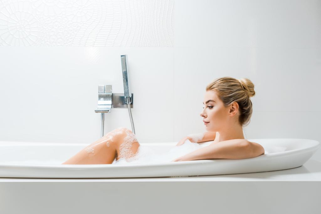 mulher bonita e loira tomando banho com espuma e olhando para longe no banheiro
  - Foto, Imagem