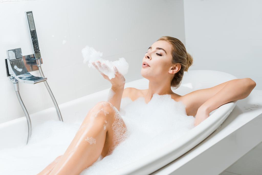 mulher atraente e loira tomando banho e brincando com espuma no banheiro
  - Foto, Imagem