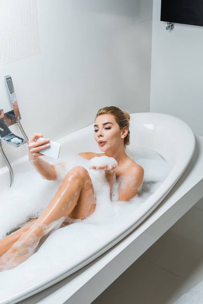 houkutteleva ja hymyilevä nainen kylvyssä, puhaltaa vaahtoa ja ottaa selfie kylpyhuoneessa
  - Valokuva, kuva