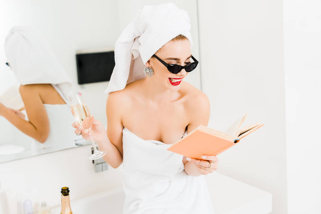 atrakcyjny i uśmiechnięta kobieta w okulary przeciwsłoneczne i ręczniki trzyma kieliszek do szampana czytanie książki  - Zdjęcie, obraz