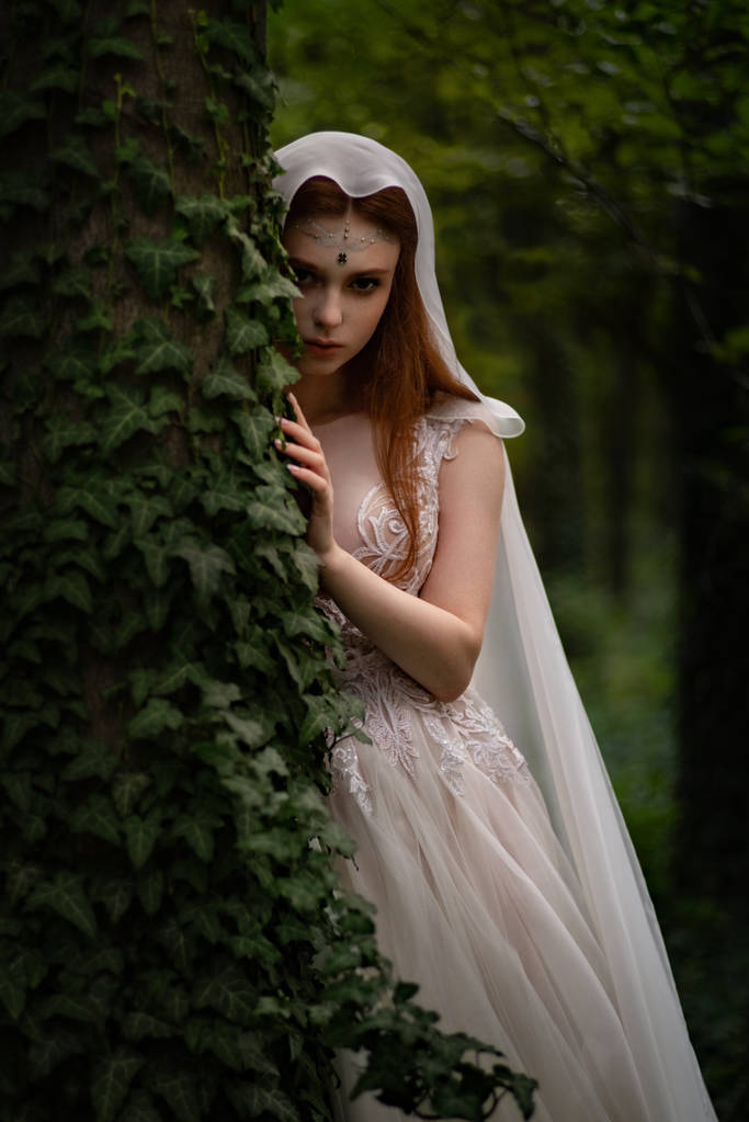atractiva mujer de jengibre elfo en vestido blanco y capa posando en jardín verde
  - Foto, Imagen