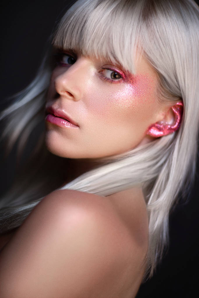 Porträt einer attraktiven blonden Frau mit glitzerndem Augen- und Ohr-Make-up, die vor dunklem Studiohintergrund in die Kamera blickt - Foto, Bild
