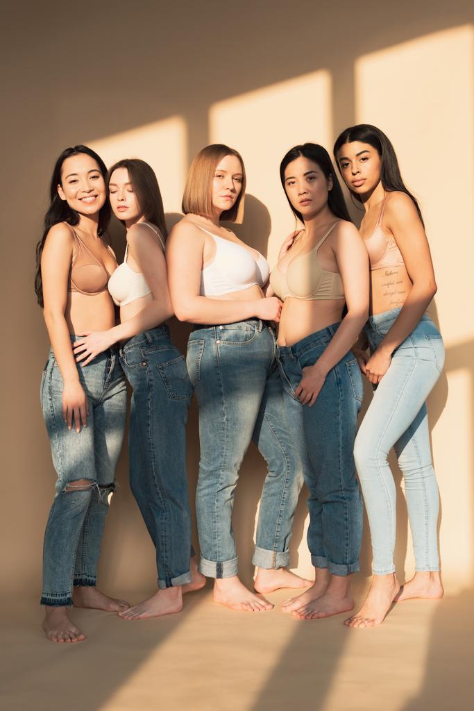 five beautiful multicultural women in blue jeans and bras standing together in sunlight, body positivity concept - Photo, Image