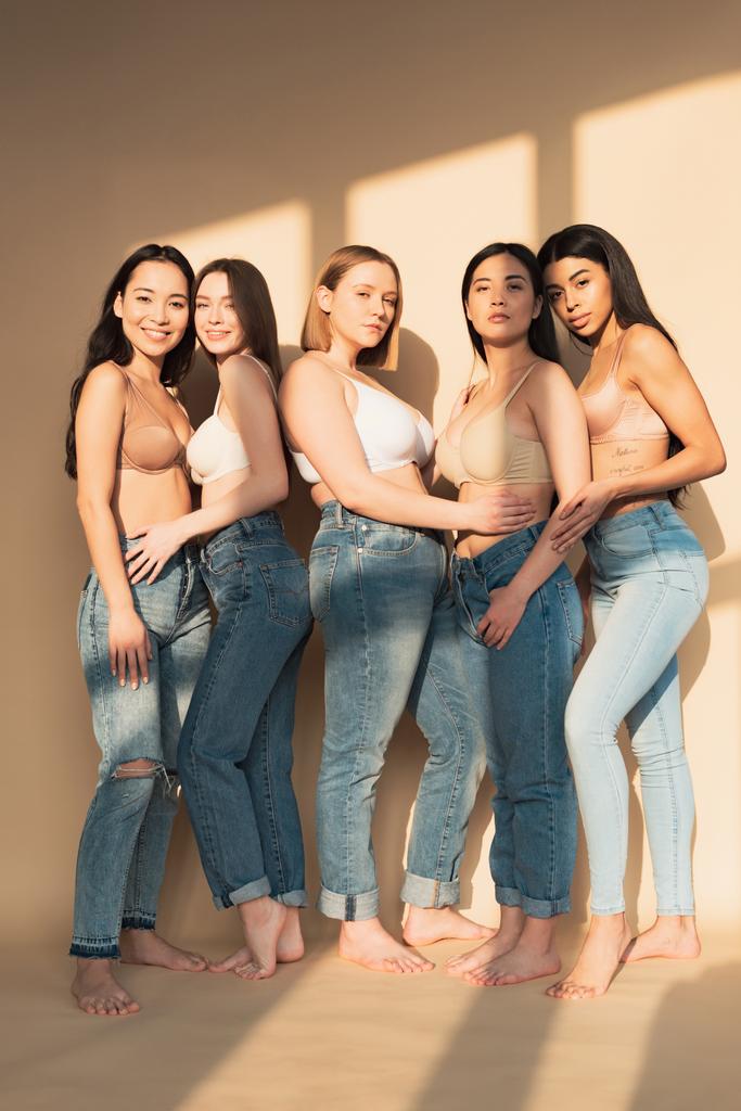 five attractive multicultural women in blue jeans and bras standing in sunlight and looking at camera, body positivity concept - Photo, Image