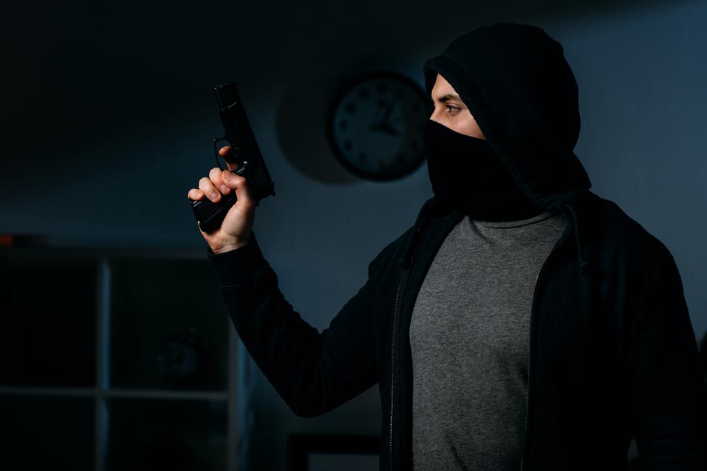 Karanlık odada tabanca tutan ve uzağa bakarak maskeli hırsız - Fotoğraf, Görsel