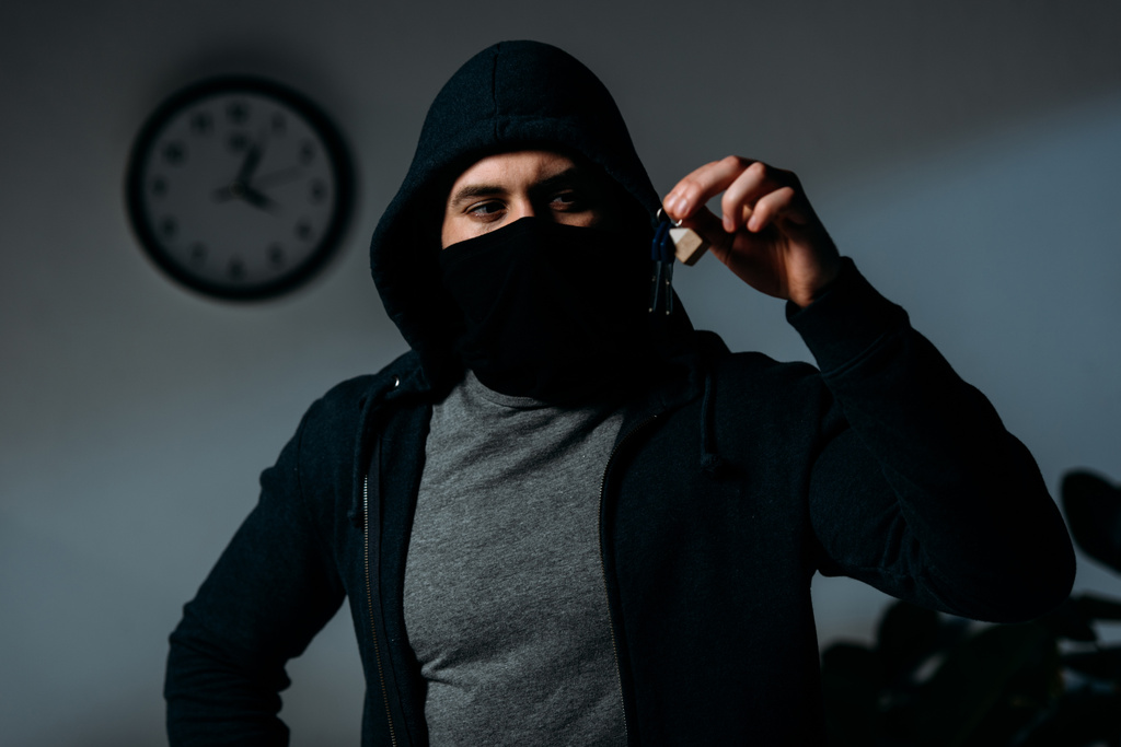 Confident thief in mask and hoodie standing in dark room and looking at keys - Photo, Image