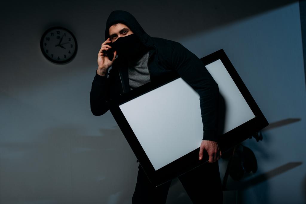 Ladrón en máscara hablando en el teléfono inteligente, mientras que el robo de televisión de pantalla plana con pantalla en blanco
 - Foto, imagen