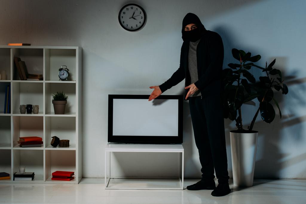 Злодій у масці та светрі, що вказує руками на телевізор з плоским екраном з порожнім екраном
 - Фото, зображення