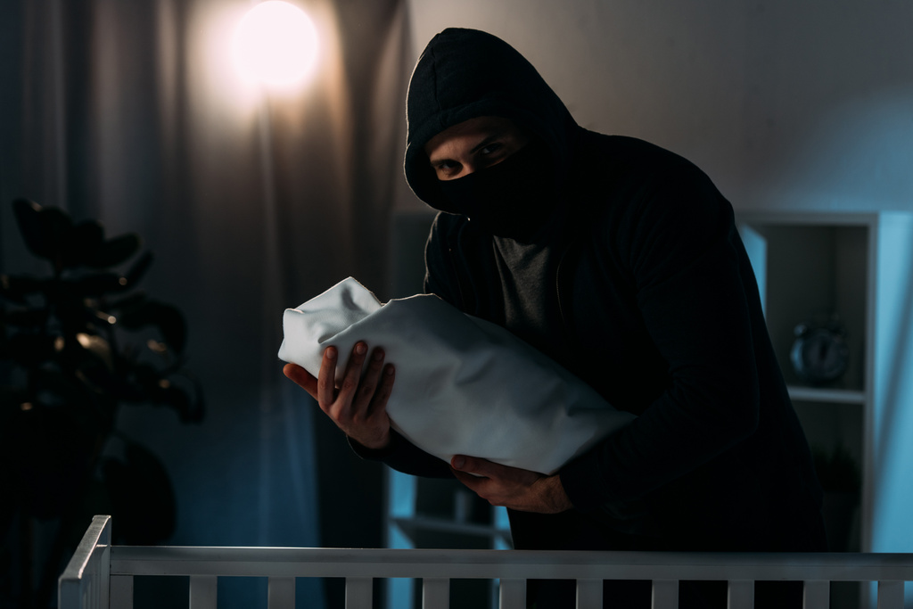 Secuestrador con máscara y sudadera con capucha que sostiene al bebé en habitación oscura
 - Foto, imagen