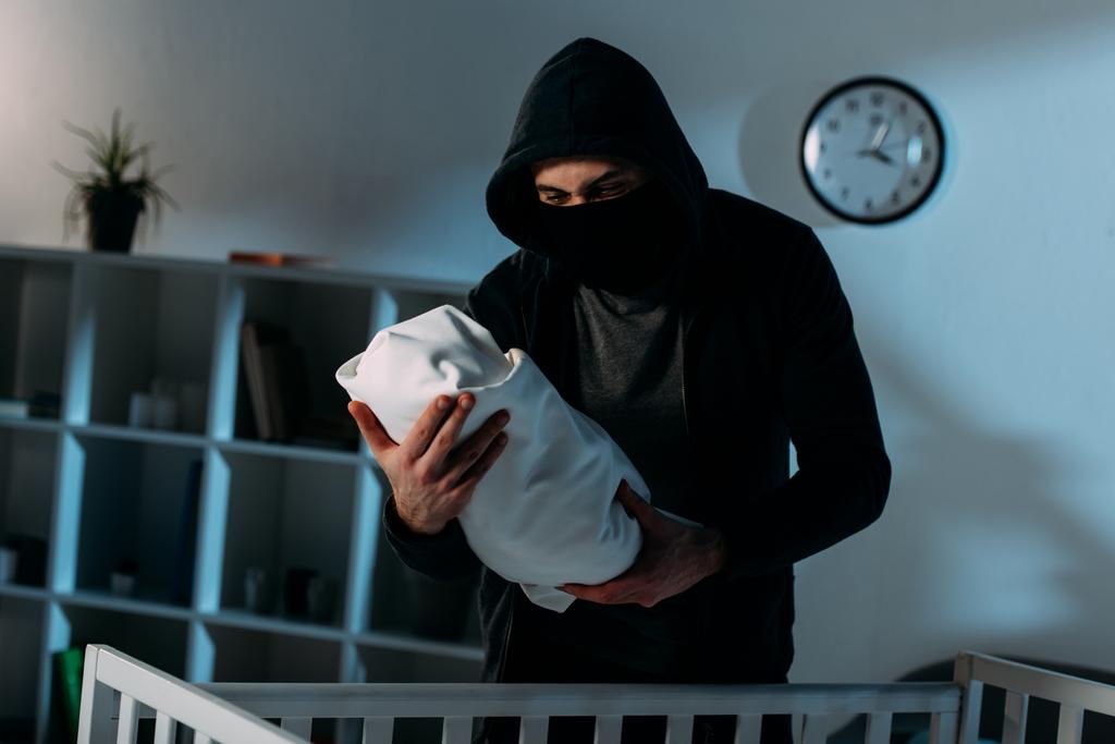 Secuestrador con máscara y sudadera con capucha negra de pie cerca de la cuna y sosteniendo al bebé
 - Foto, imagen