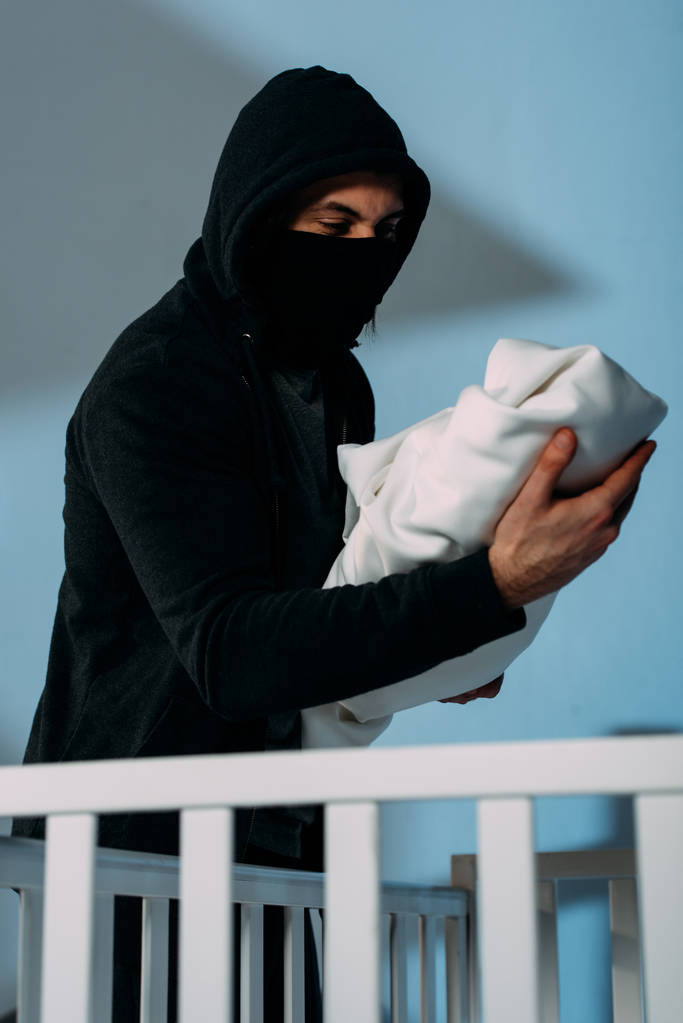 Kidnapper in mask standing beside crib and holding infant child - Photo, Image