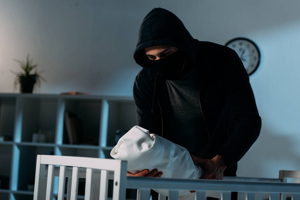 Secuestrador con máscara y sudadera con capucha de pie cerca de la cuna y sosteniendo al bebé
 - Foto, imagen