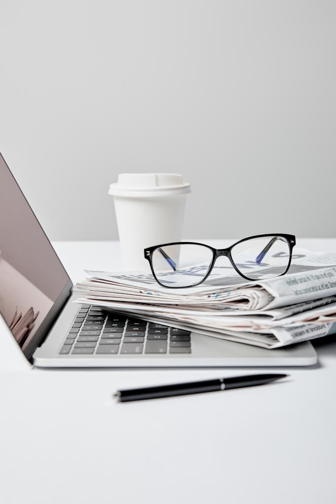 Laptop mit leerem Bildschirm in der Nähe von Zeitungen, Brille, Stift und Pappbecher auf grau  - Foto, Bild