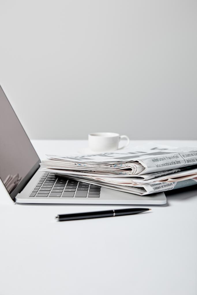 messa a fuoco selettiva del computer portatile con schermo bianco vicino a giornali, penna e tazza su grigio
  - Foto, immagini