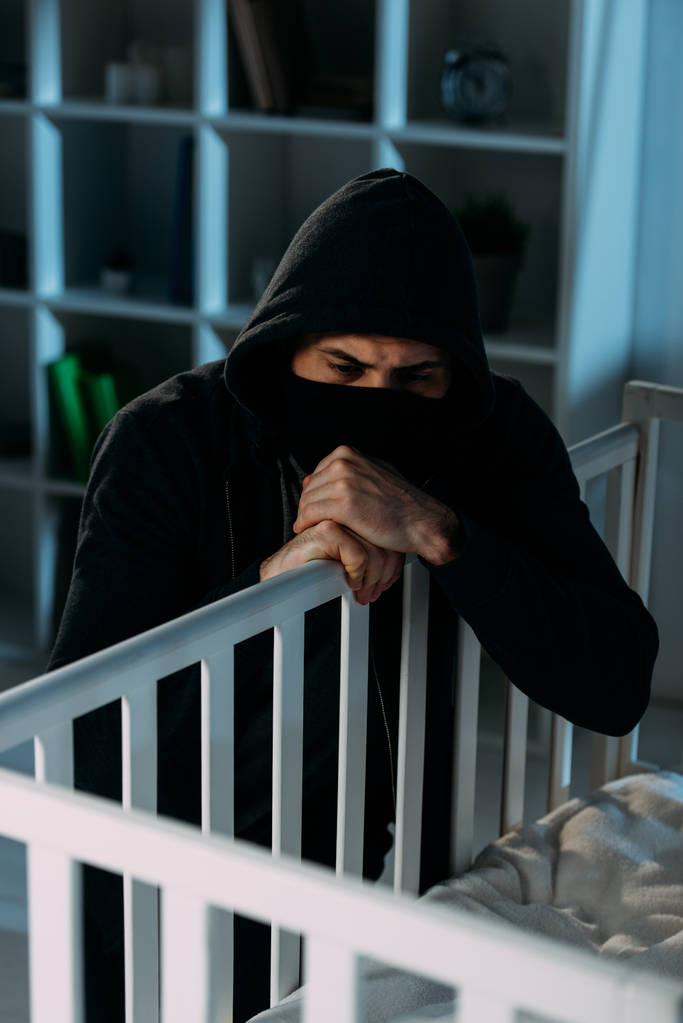 Secuestrador pensativo con máscara negra buscando en cuna en habitación oscura
 - Foto, Imagen