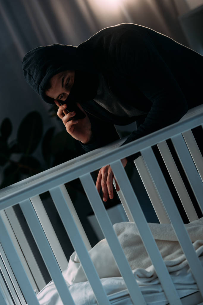 Secuestrador en máscara hablando en el teléfono inteligente mientras está cerca de la cuna
 - Foto, Imagen