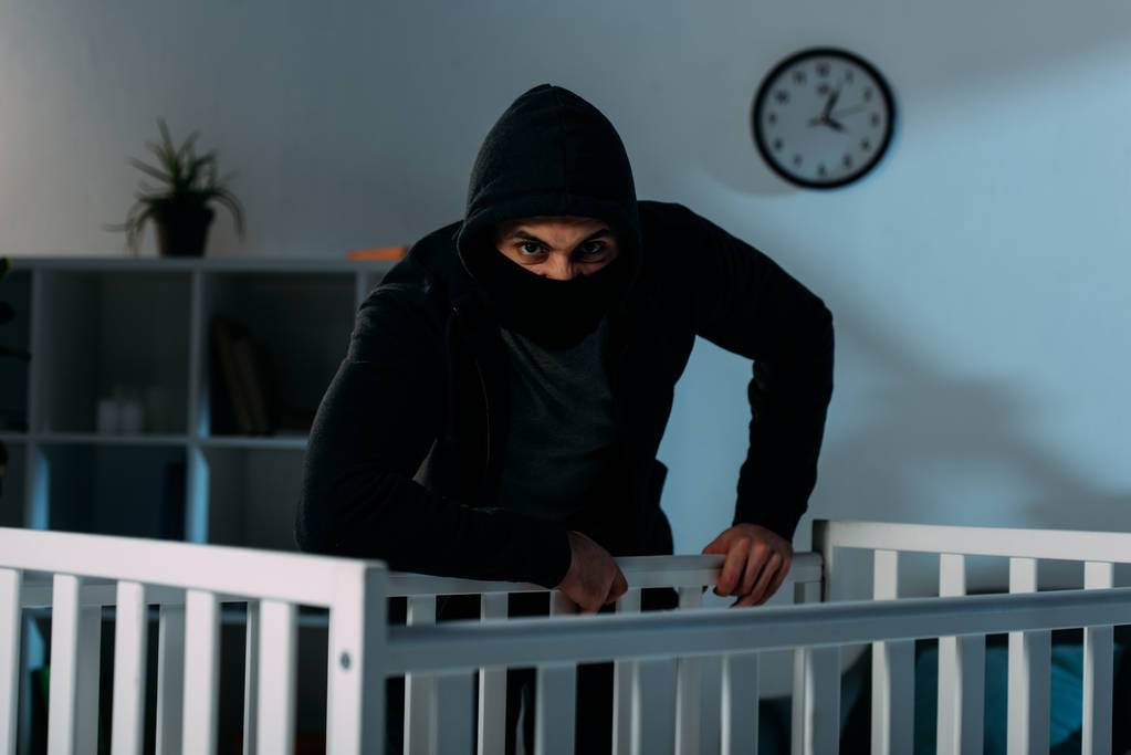 Злой похититель в черной маске стоит возле кроватки и смотрит в камеру
 - Фото, изображение