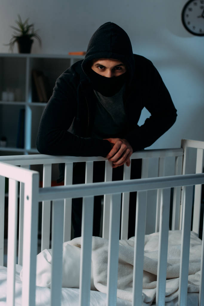 Criminal en máscara y sudadera con capucha de pie cerca de cuna en habitación oscura
 - Foto, imagen