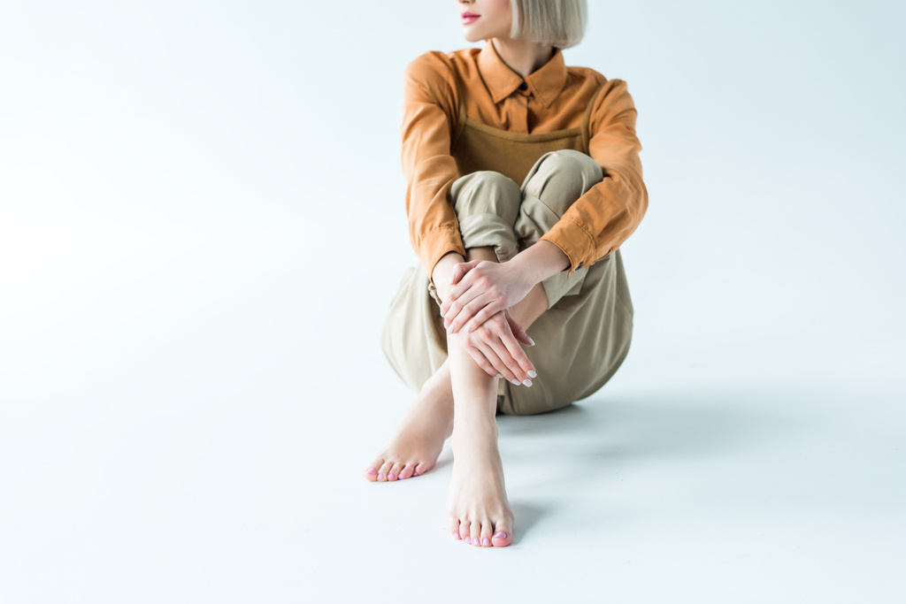 обрезанный вид стильной молодой женщины, сидящей со скрещенными ногами на белом
 - Фото, изображение