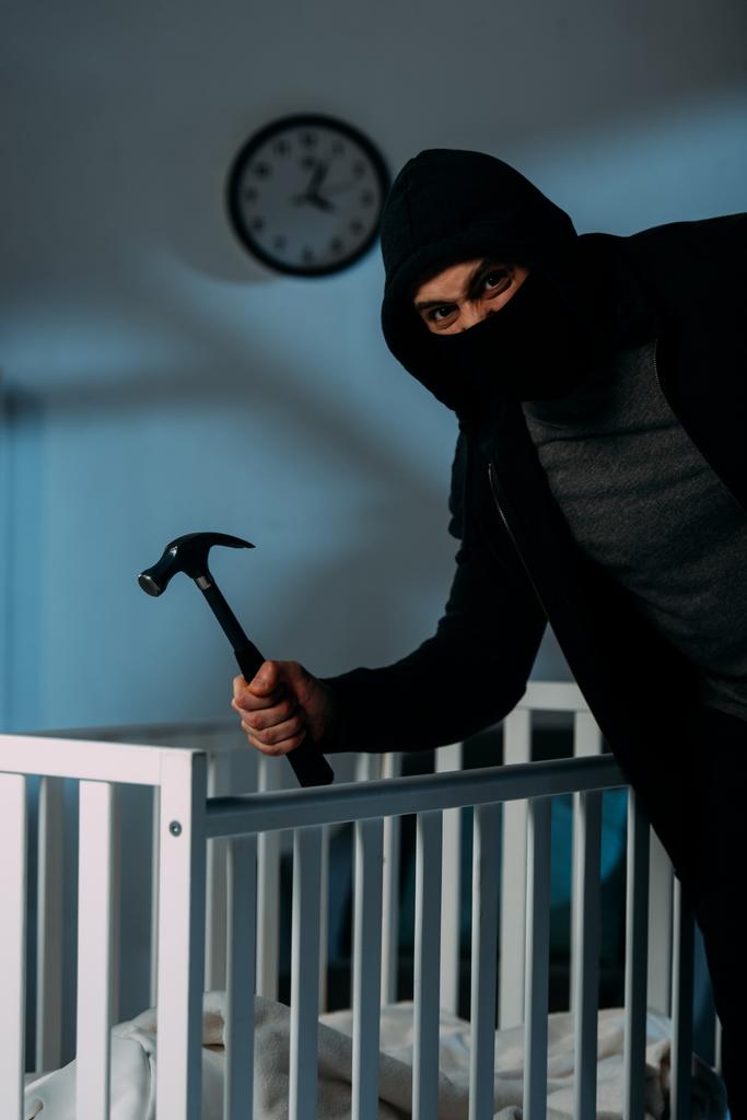 Angry criminal in mask standing near crib and holding hammer - Photo, Image