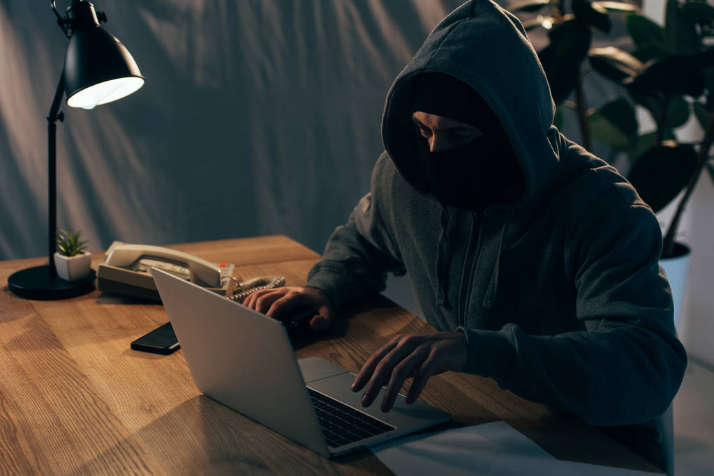 Criminal en máscara y sudadera con capucha escribiendo en el teclado del ordenador portátil en habitación oscura
 - Foto, Imagen