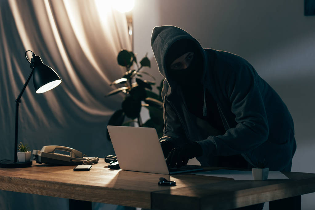 パーカーと暗い部屋でノート パソコンを使用してマスクの犯罪者 ロイヤリティフリー写真 画像素材