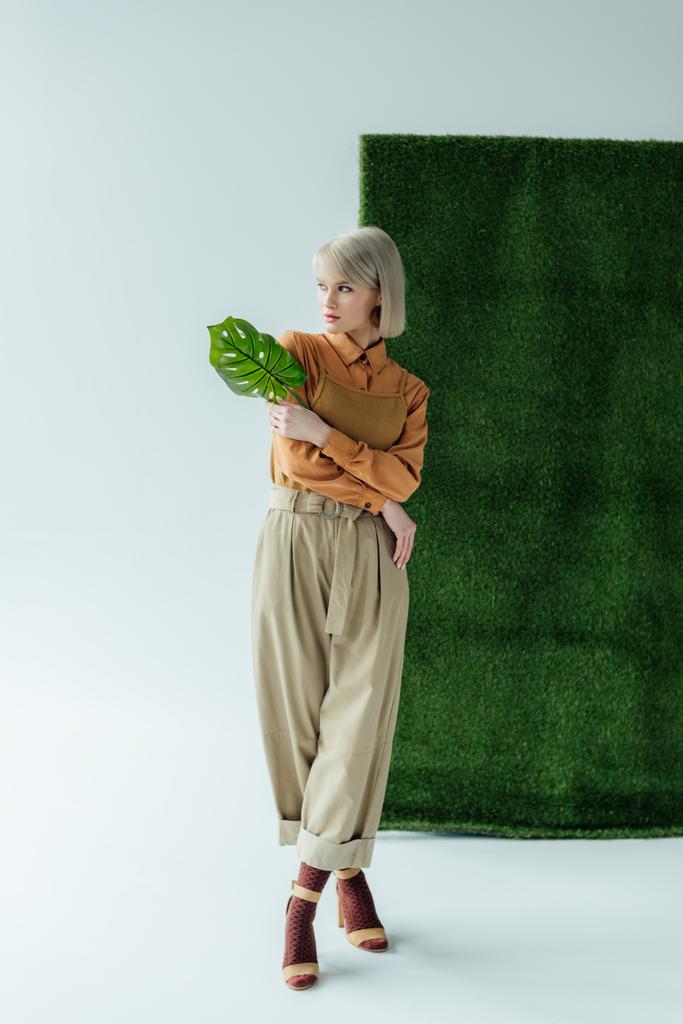 schöne junge Frau hält Monstera Blatt, während sie auf weiß mit grünem Gras posiert  - Foto, Bild