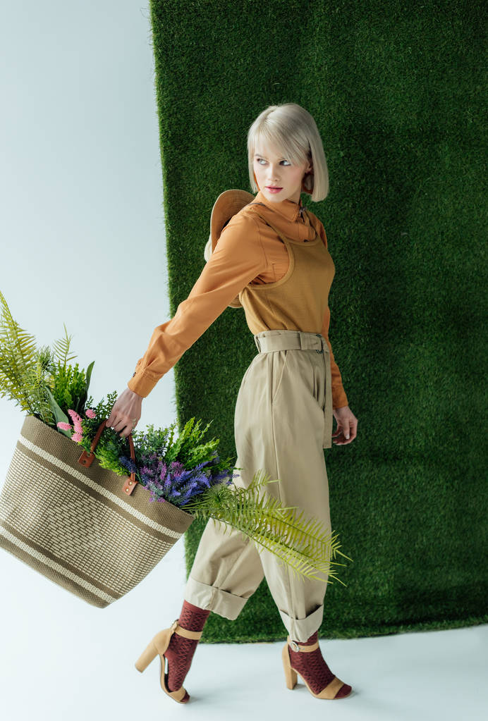 hermosa mujer joven con estilo sosteniendo bolsa con helecho y flores en blanco con hierba verde
  - Foto, imagen