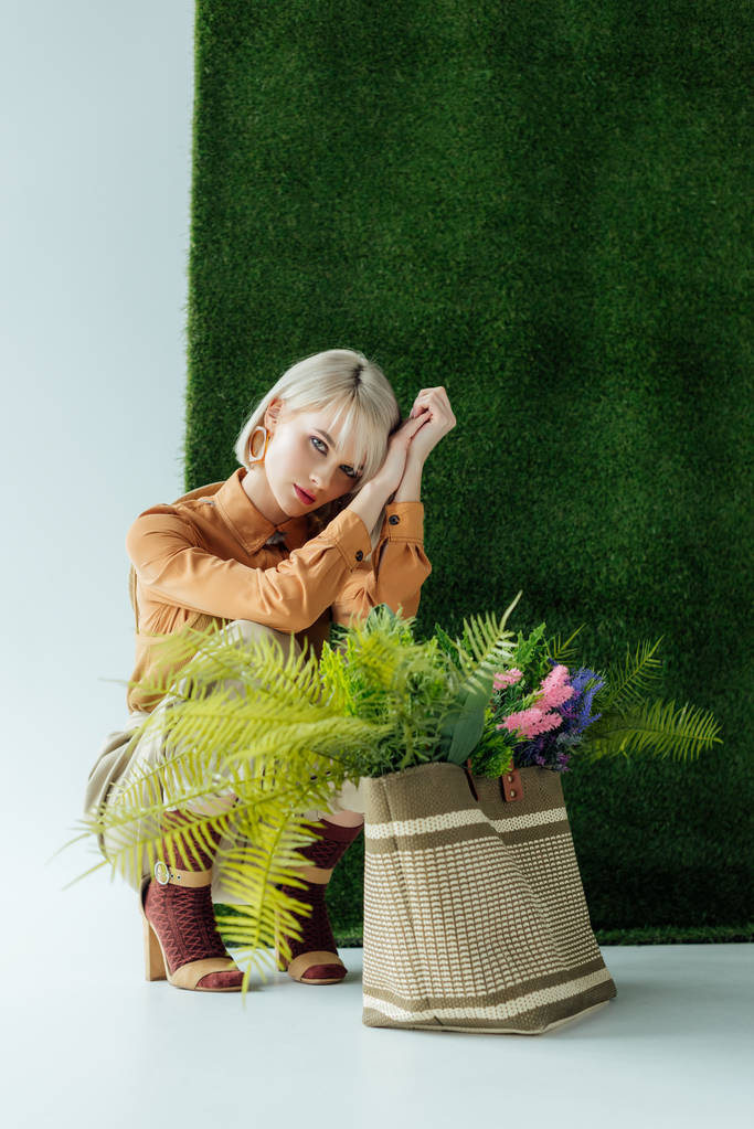 mooie modieuze meisje poseren in de buurt van tas met varens en bloemen op wit met groen gras  - Foto, afbeelding