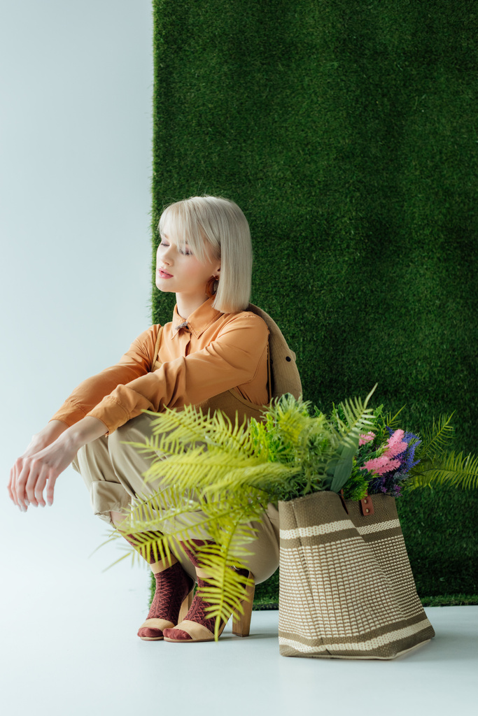 piękna stylowa dziewczyna stwarzających w pobliżu torba z paproci i kwiaty na białym tle z zielonej trawie  - Zdjęcie, obraz