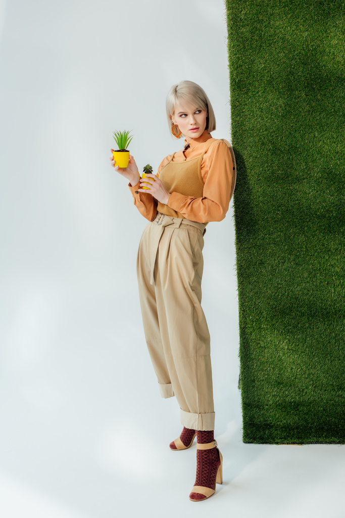 mooie stijlvolle blond meisje houdt van bloempotten op grijs met groen gras  - Foto, afbeelding